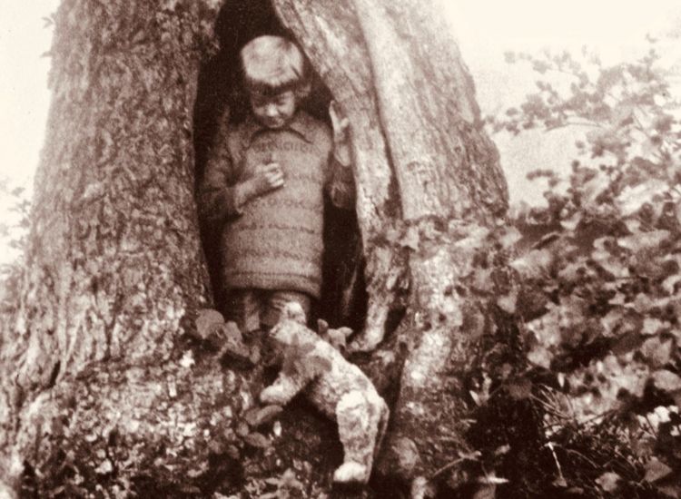 An old photo of a boy holding a teddy bear, leaning against a tree